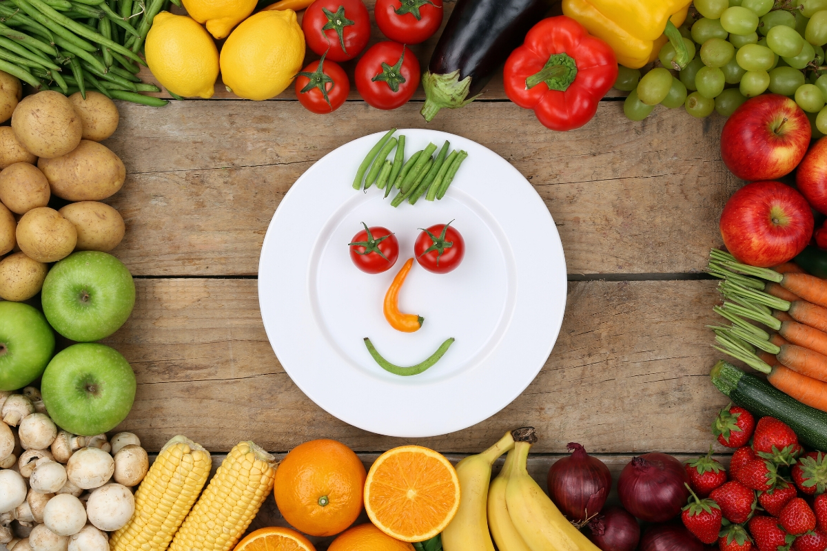 A plate with a funny face made of vegetables surrounded by other fruits and vegetables.
