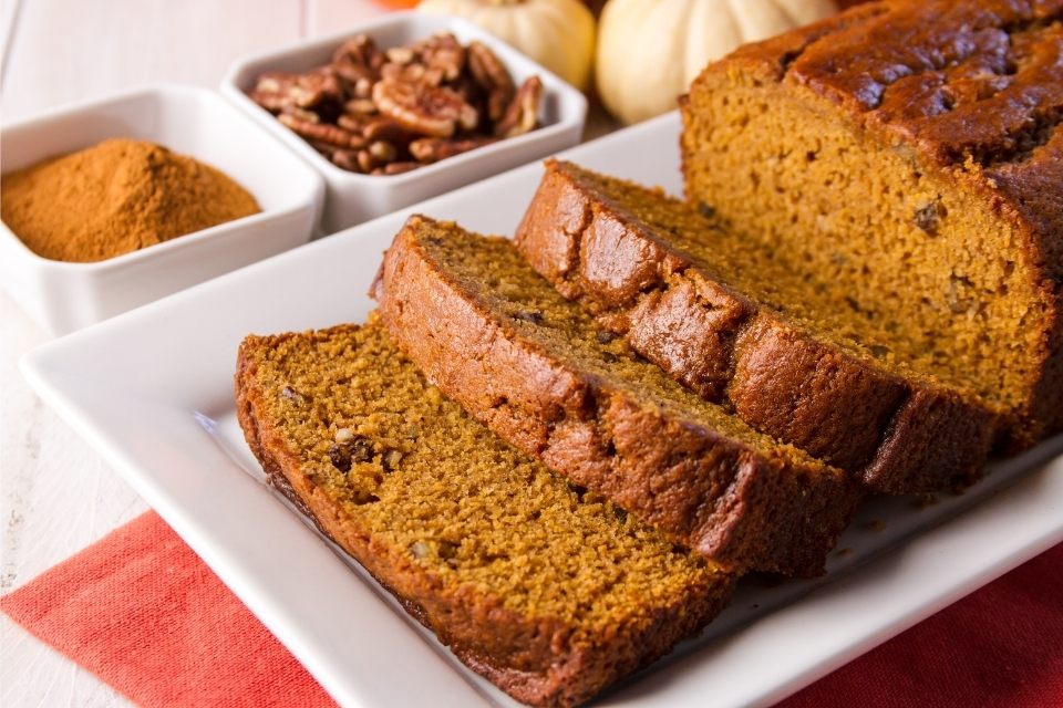 Pumpkin bread with pecans on a serving plate with some slices already cut. Spices and pecans on recipients on the side.