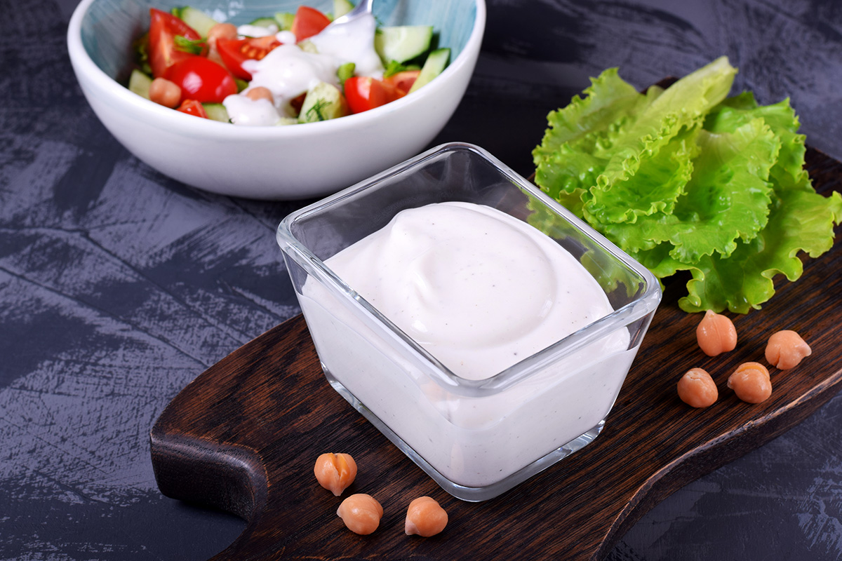 A container with vegan mayonnaise made with aquafaba on the foreground, and a salad dressed with this mayo in the background.