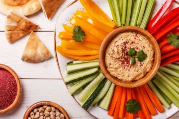 Hummus en un cuenco rodeado de verduras crudas cortadas en bastoncitos.