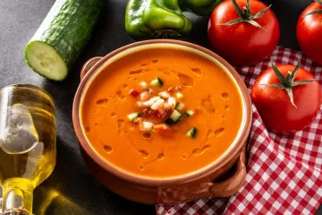 Un cuenco de gazpacho con sus guarniciones típicas rodeado de los ingredientes que se usan para hacerlo.