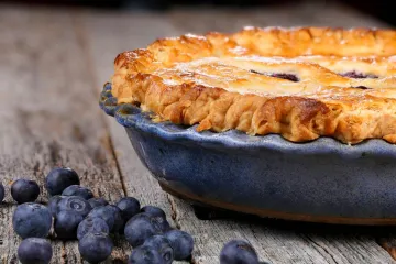 Tarta americana de arándanos azules en una fuente de cerámica azul, con corteza dorada y arándanos frescos sobre una mesa de madera rústica.