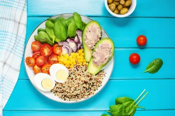Plato completo con atún, aguacate relleno, huevo duro, tomates cherry, cebolla morada, maíz, espinacas y una mezcla de arroz y quinoa, sobre una mesa de madera azul.