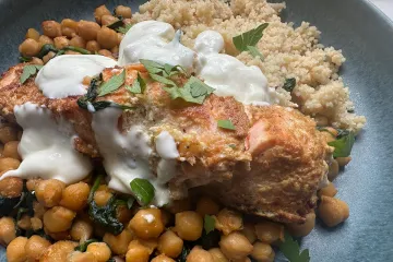 Lomo de salmón especiado y dorado, napado con salsa de yogur y espolvoreado de perejil, sobre garbanzos salteados con espinacas y con cuscús al lado, en un plato azul.