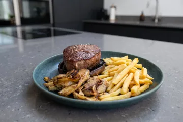 Sobre la encimera de la cocina, un plato con un tournedós sobre una cabeza de seta Porotbello acompañado de aclcachofas y patatas paja.