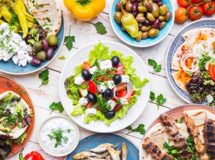 Una selección de platos de la cocina griega, incluyendo meze, gyros, souvlaki, pan pita, ensalada griega, tzatziki, queso feta, aceitunas.
