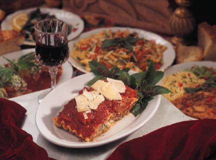 Mesa italiana con lasaña, otros platos de pasta, y una copa de vino tinto.