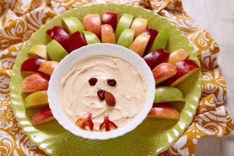 Un cuenco con crema de queso al caramelo, decorado como un pollito, rodeado de trozos de manzana.