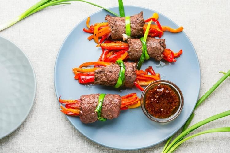 Un plato con varios rollitos de carne rellenos de tiras de pimiento y atados con hojas de cebolleta.