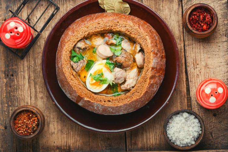 Zurek, una sopa polaca tradicional, servida en un bol de pan comestible, con salchicha, huevos duros y decorada con perejil fresco, con elementos rústicos y condimentos alrededor del bol.
