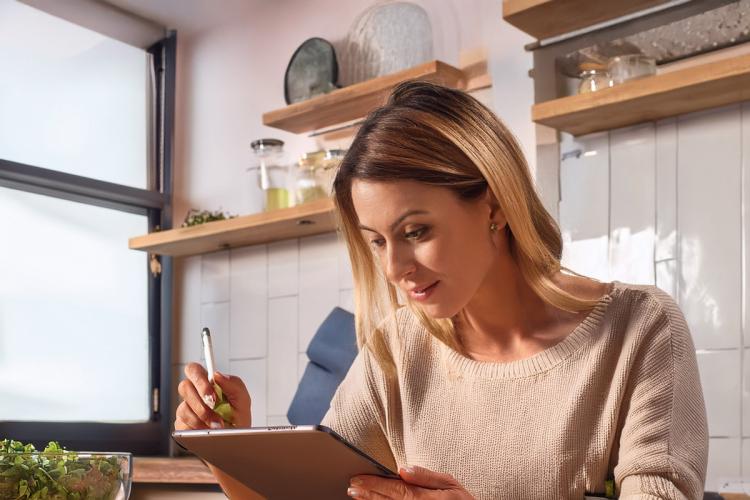 Una mujer sentada delante de la mesa de la cocina tomando notas sobre la receta que ha preparado en una tableta electrónica.