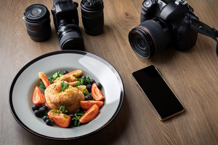 Un plato de comida sobre una mesa, a su lado una cámara fotográfica, varios objetivos y un teléfono móvil.