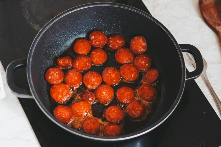 Trozos de salchicha húngara roja al pimentón friéndose en una sartén negra de dos asas.