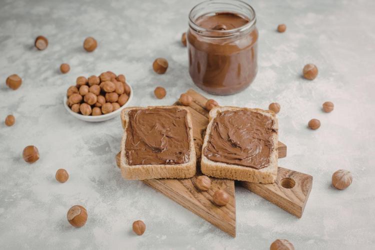 Dos tostadas untadas con pasta de chocolate y avellana con un tarro de vidrio lleno de pasta detrás. Están rodeados de avellanas peladas.