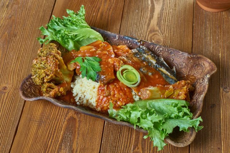 Plato de cou cou con pez volador en salsa de tomate, acompañado de brócoli, puerro y hojas verdes, servido en una fuente de cerámica sobre una mesa de madera.
