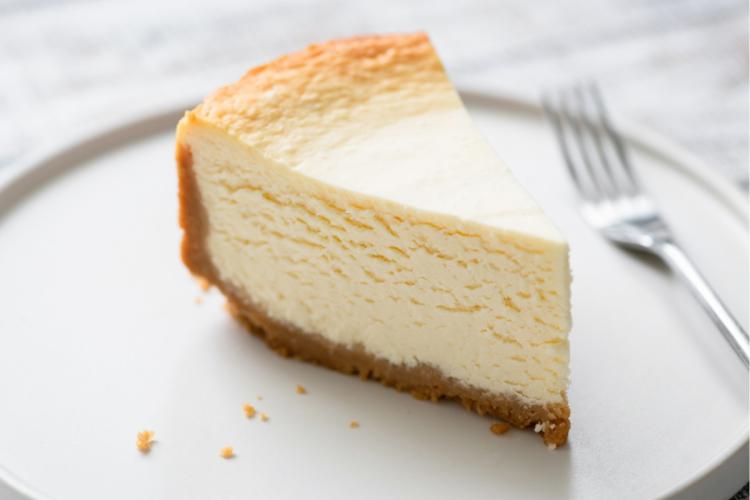 Porción de tarta de queso clásica con base de galleta servida en un plato blanco, con un tenedor desenfocado al fondo.