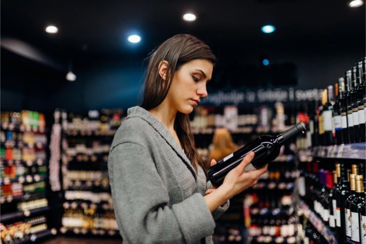 Mujer joven leyendo la etiqueta de una botella de vino tinto en el pasillo de una tienda de vinos.