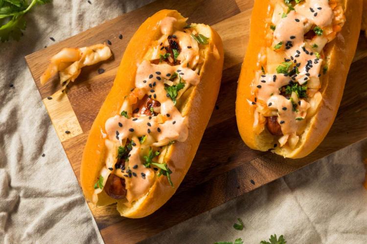 Dos perritos calientes de estilo coreano en pan brioche, cubiertos con kimchi, mayonesa picante y semillas de sésamo negro, sobre una tabla de madera.