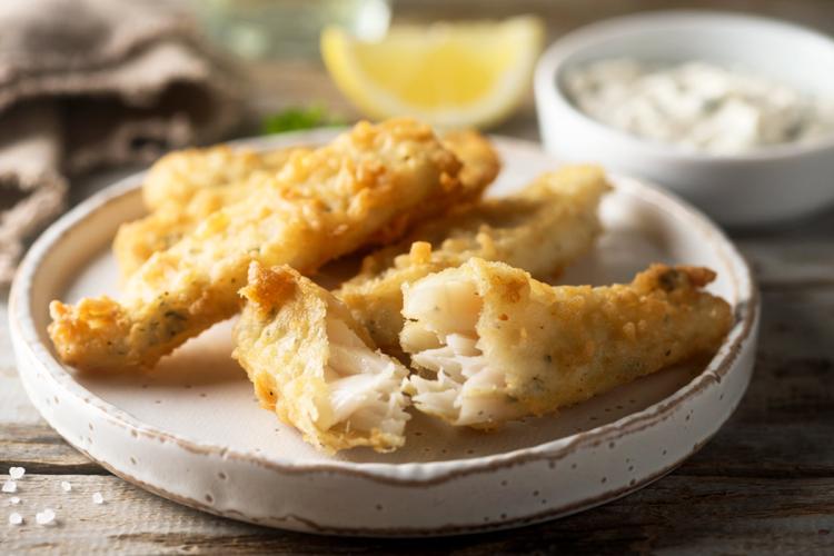 Varitas de pescado rebozadas y fritas, doradas y crujientes, sobre un plato blanco con un limón y una salsa tártara al fondo - imagen sintética.