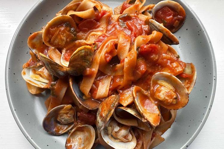 Plato hondo gris con cintas de pasta (tagliatelle) en salsa de tomate marinera rodeadas de almejas abiertas con sus valvas, sobre una mesa blanca.