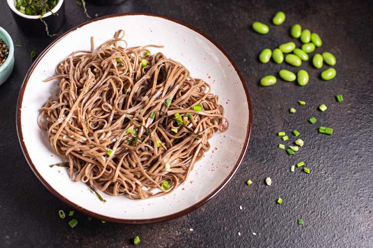 Cuenco blanco con fideos soba fríos aliñados, espolvoreados con cebolleta picada y hebras de alga, junto a granos de edamame y sésamo esparcidos sobre una superficie oscura.
