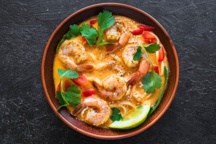 Cuenco de barro con laksa de gambas en un caldo cremoso anaranjado, con fideos, tiras de pimiento rojo, hojas de cilantro, semillas de sésamo y una rodaja de lima, visto desde arriba sobre una superficie oscura.