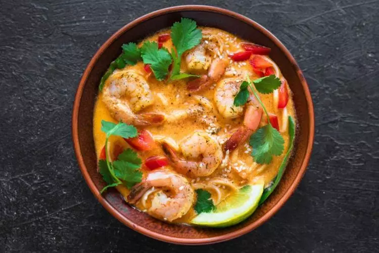 Cuenco de barro con laksa de gambas en un caldo cremoso anaranjado, con fideos, tiras de pimiento rojo, hojas de cilantro, semillas de sésamo y una rodaja de lima, visto desde arriba sobre una superficie oscura.