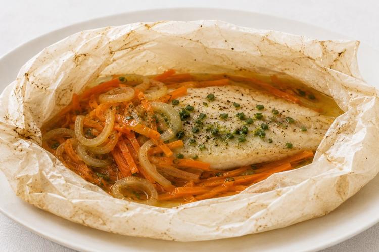 Filete de pescado blanco cocinado en papillote sobre juliana de zanahoria y aros de cebolla, con cebollino picado por encima, dentro del papel de horno abierto sobre un plato blanco.