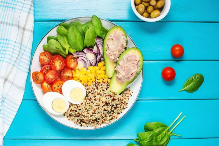 Plato completo con atún, aguacate relleno, huevo duro, tomates cherry, cebolla morada, maíz, espinacas y una mezcla de arroz y quinoa, sobre una mesa de madera azul.