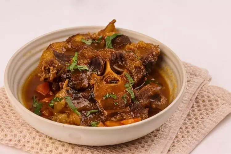 Cuenco de cerámica clara con rabo de toro braseado en una salsa espesa y brillante, con trozos de zanahoria y perejil picado por encima, sobre un paño de cocina beige.