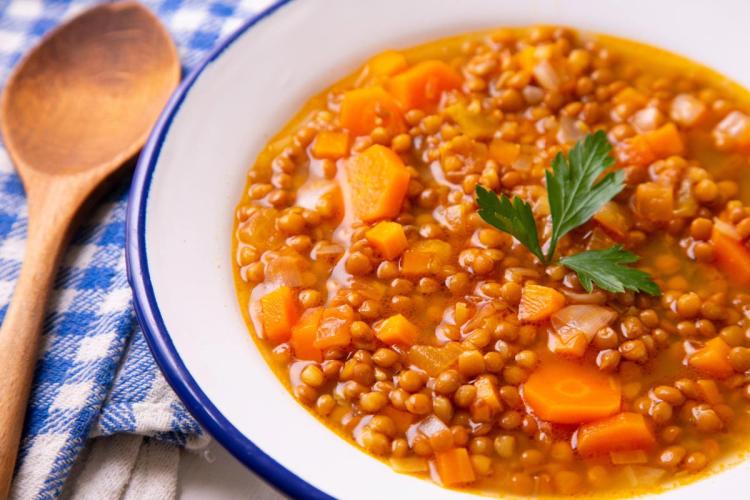 Cuenco blanco lleno de sopa de lentejas con zanahoria en dados, decorado con una hoja de perejil, junto a una cuchara de madera sobre un paño de cuadros azules.