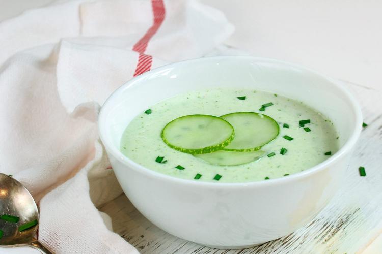 Cuenco blanco con sopa fría de pepino y calabacín, decorada con rodajas de pepino y cebollino picado, sobre una mesa de madera clara.