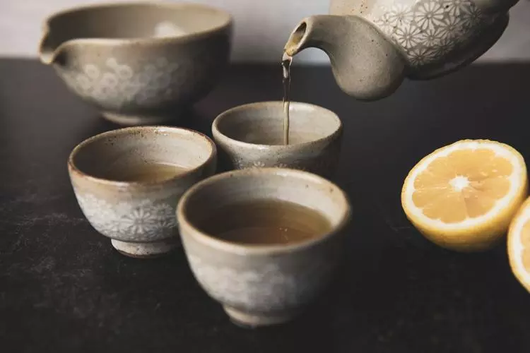 Té de color ámbar cayendo desde una tetera de gres en unas tazas pequeñas de cerámica con motivos florales blancos, sobre una superficie de piedra oscura y junto a un limón partido por la mitad.