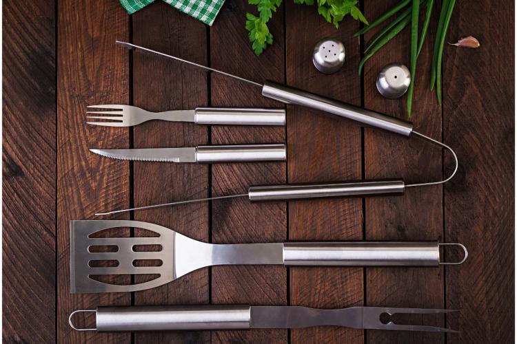 Juego de herramientas de barbacoa de acero inoxidable —tenedor, cuchillo, pinchos, espátula y pinzas— dispuestas sobre una mesa de madera oscura, junto a hierbas frescas, ajo y saleros.