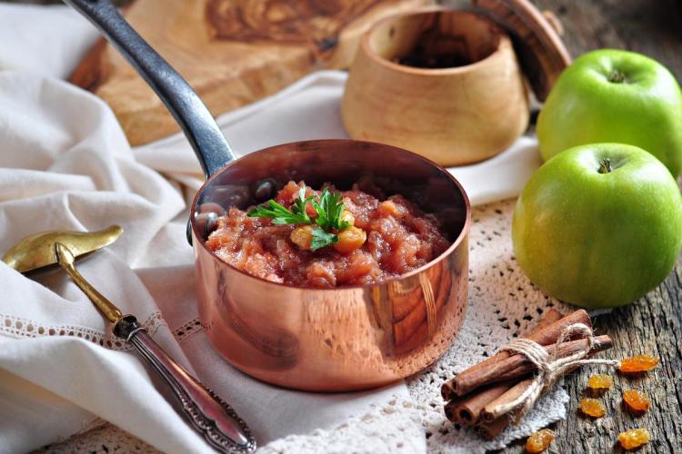 Un cazo de cobre con chutney de manzana rodeado de algunos de sus ingredientes.