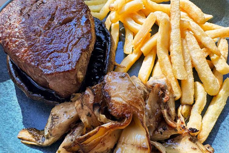 Solomillo de ternera a la plancha sobre un sombrero de portobello, acompañado de setas y alcachofas salteadas y de patatas fritas, en un plato azul.