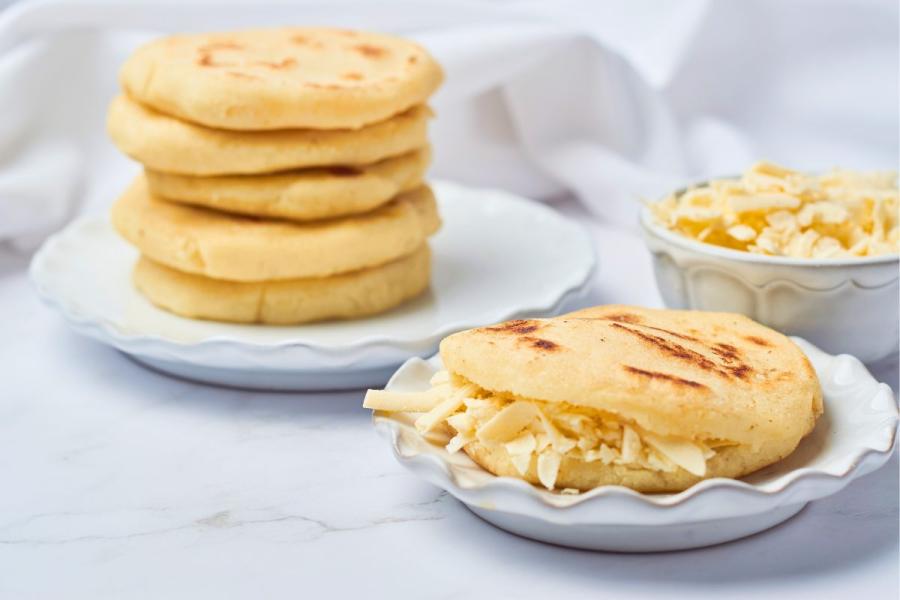 Una arepa rellena de queso rallado grueso, con un plato con una pila de arepas y un cuenco con queso al lado.