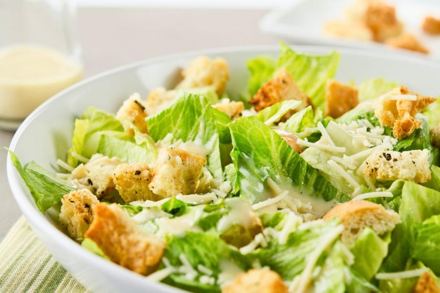 Una ensalada César en su ensaladera con un poco de aliño César en un recipiente al fondo.