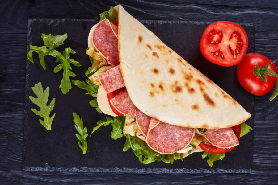 Una piadina en bocadillo con rodajas de salami, queso mozzarella, tomate, calabacín asado y rúcula.