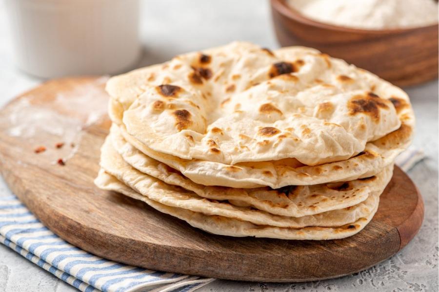 Una pila de panes planos tipo chapati recién hechos, colocados sobre una tabla de madera con un paño rayado debajo.