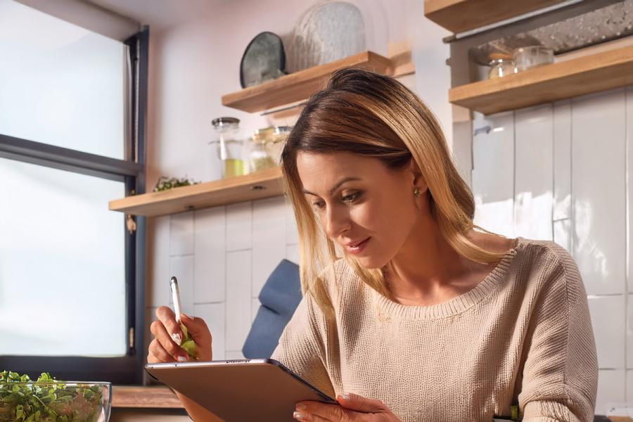 Una mujer sentada delante de la mesa de la cocina tomando notas sobre la receta que ha preparado en una tableta electrónica.