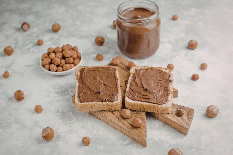 Dos tostadas untadas con pasta de chocolate y avellana con un tarro de vidrio lleno de pasta detrás. Están rodeados de avellanas peladas.