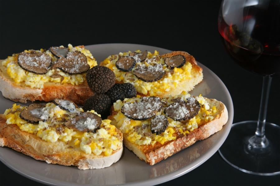 Cuatro tostadas con huevos revueltos y rodajas de trufa, servidas en un plato gris, acompañadas de una copa de vino tinto.