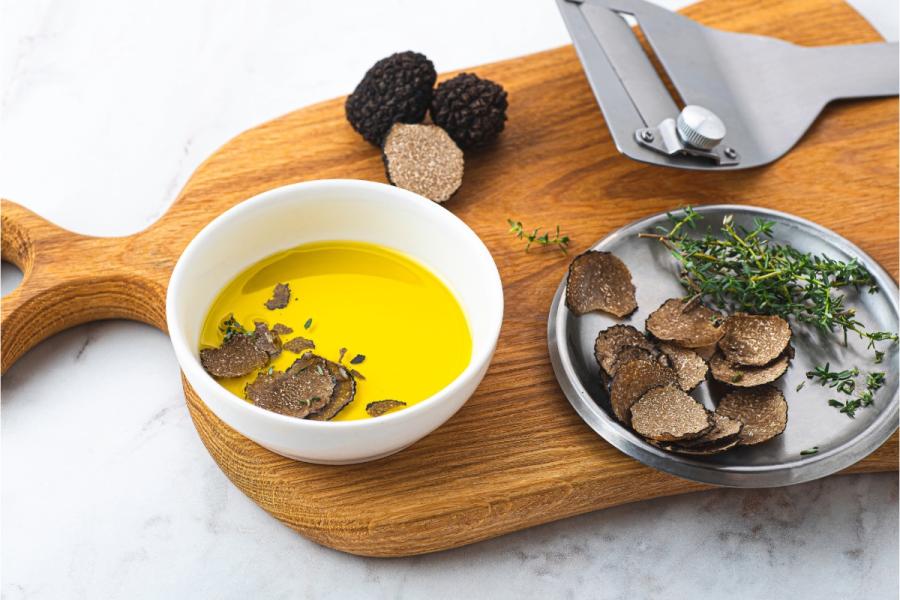 Aceite de oliva en un tazón blanco con rodajas de trufa negra y tomillo en una tabla de cortar, junto a un rallador de trufas.