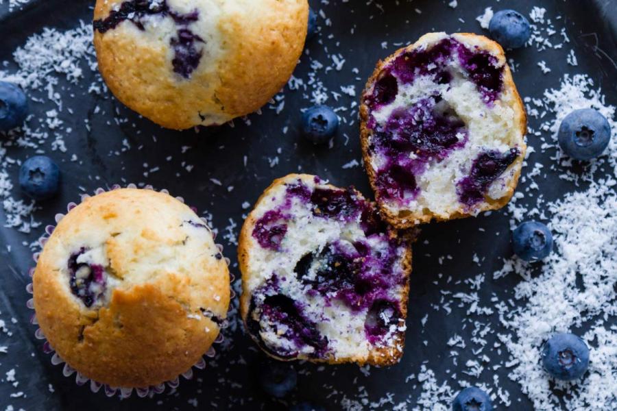Muffins de arándanos azules sobre una superficie oscura, uno de ellos partido por la mitad mostrando su interior esponjoso con abundantes arándanos, rodeados de frutos frescos y coco rallado.
