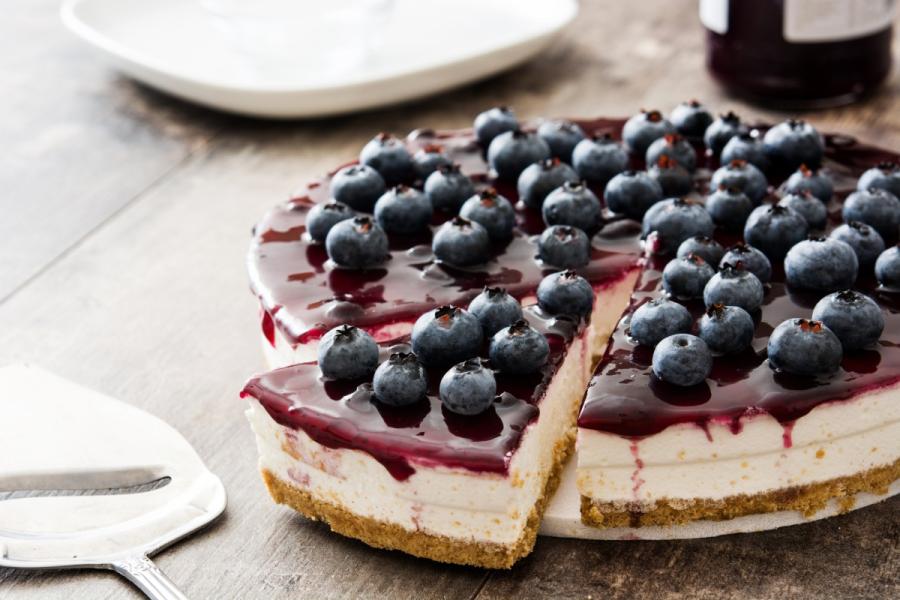 Tarta de queso cubierta con salsa de arándanos azules y arándanos frescos, con una porción cortada que muestra el relleno cremoso.