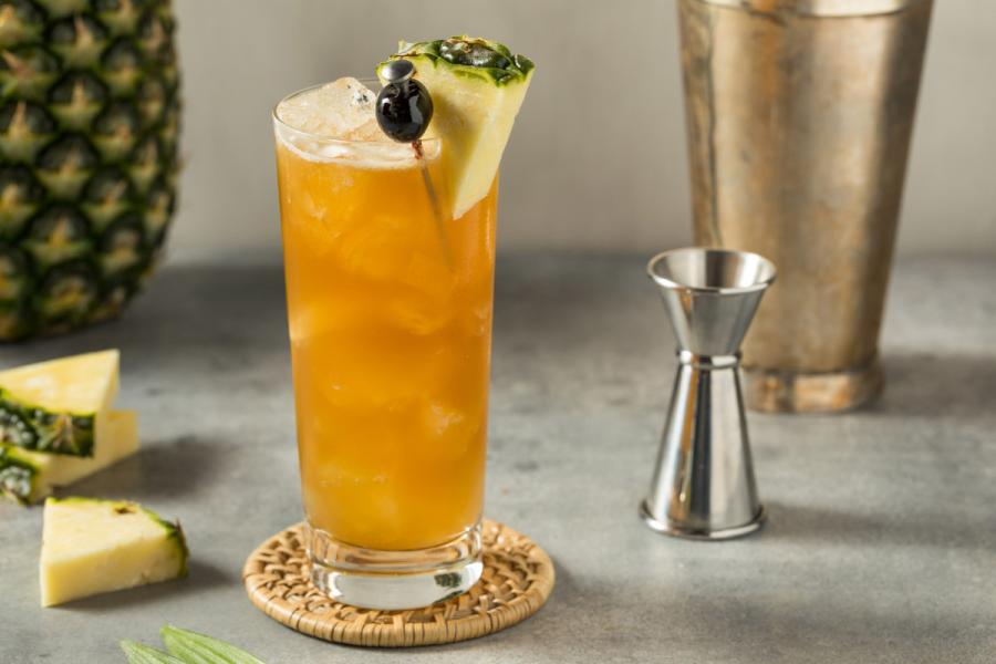 Cóctel Bahama mama servido con hielo en un vaso alto, decorado con una cuña de piña y una cereza, sobre una encimera moderna junto a utensilios de coctelería y trozos de piña fresca.