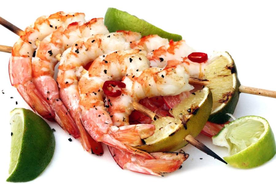 Brochetas de gambas a la parrilla con rodajas de lima asada, gajos de lima fresca y chile rojo sobre un plato blanco.