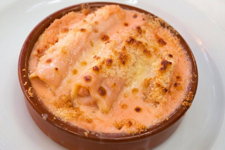 Una porción de canelones de verduras gratinados con bechamel de tomate en una cazuela de barro sobre un plato blanco.