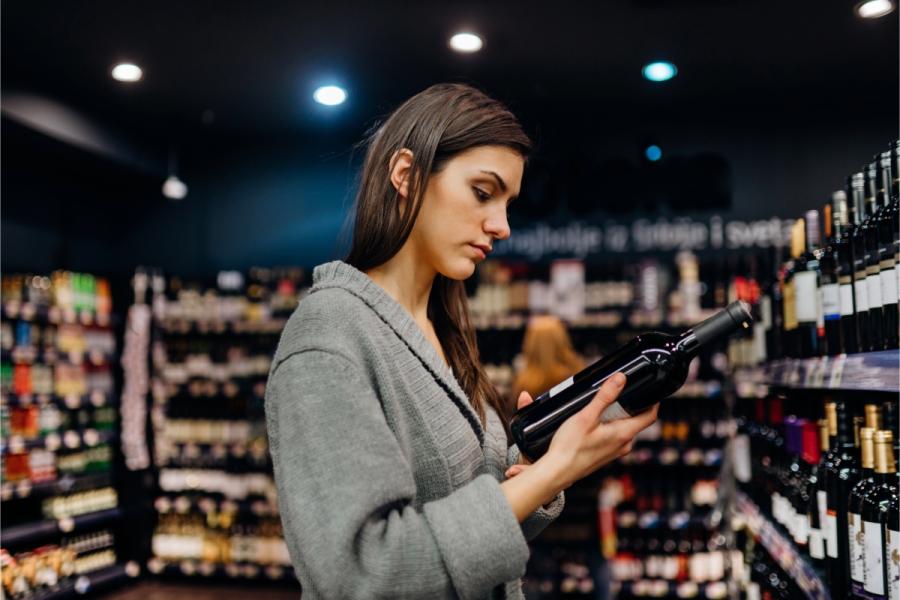 Mujer joven leyendo la etiqueta de una botella de vino tinto en el pasillo de una tienda de vinos.